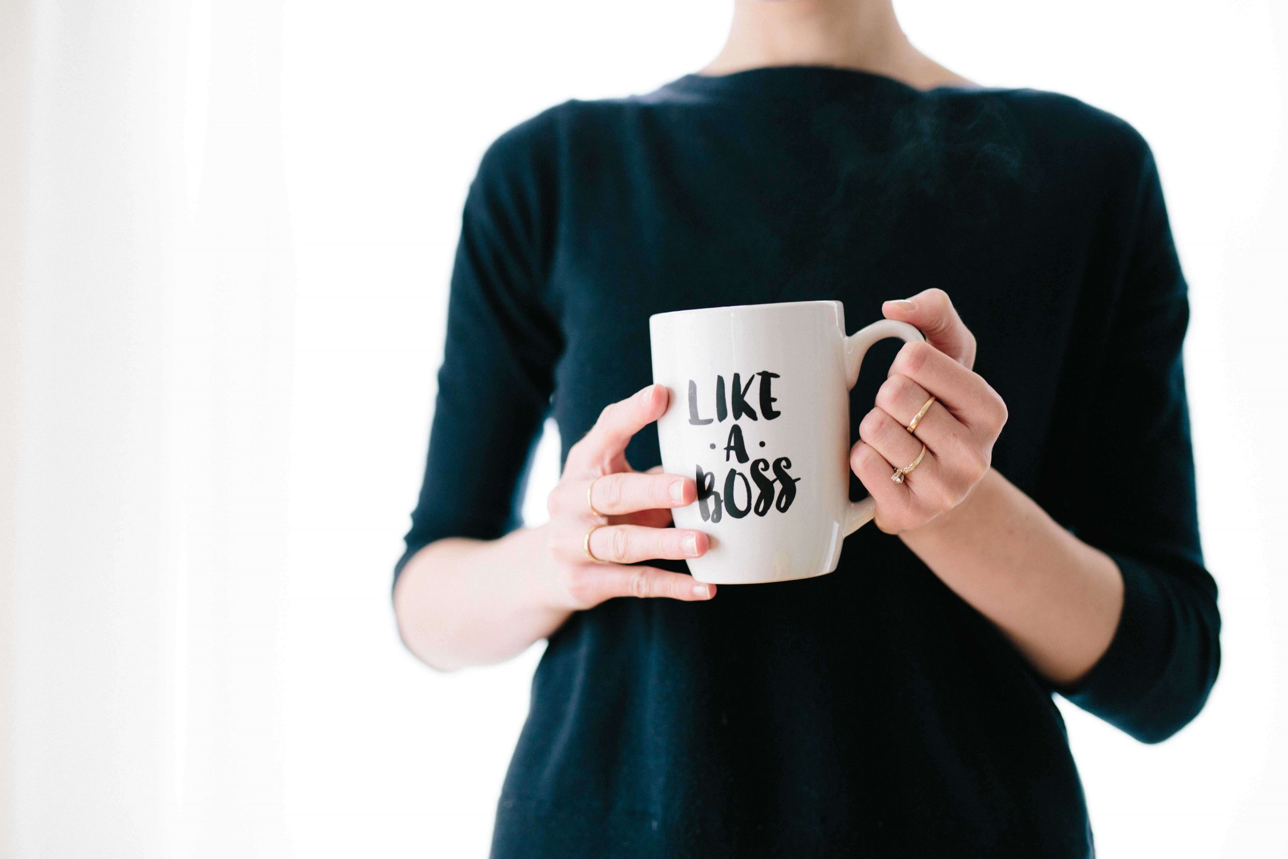 A woman's torso. She's wearing a black sweater and holding a white coffee mug that says, "Like a Boss."