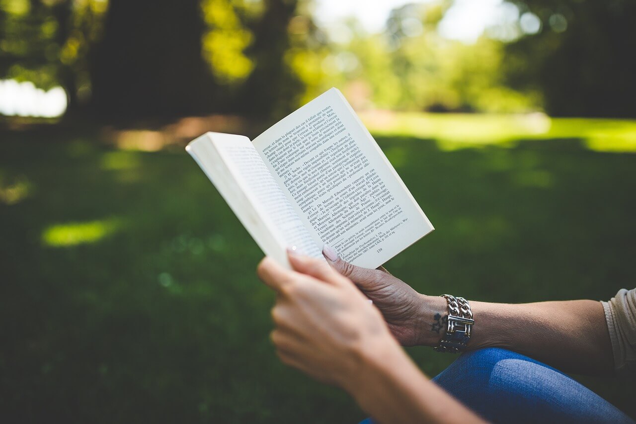 A woman holding a book in her hand, reading outside with grass and trees in the background